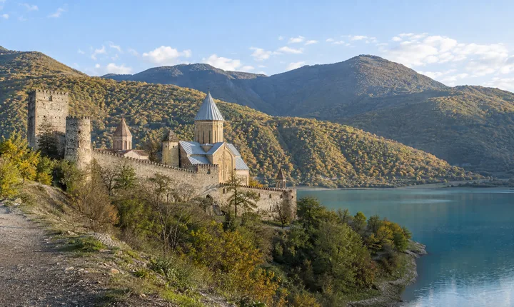 A stone fortress and domed church beside a turquoise reservoir and wooded hills A stone fortress and domed church beside a turquoise reservoir and wooded hills