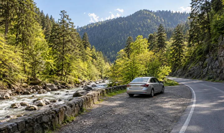 A silver sedan at a pull-off beside a paved road through a wooded Georgian river valley A silver sedan at a pull-off beside a paved road through a wooded Georgian river valley
