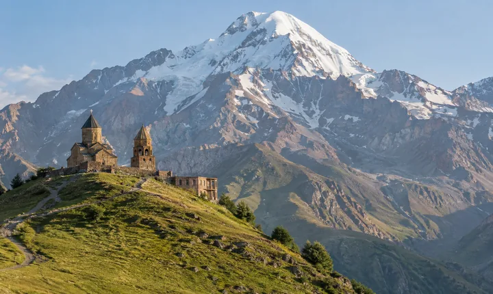 A stone church and bell tower stand on a grassy ridge below snow-covered Mount Kazbek A stone church and bell tower stand on a grassy ridge below snow-covered Mount Kazbek