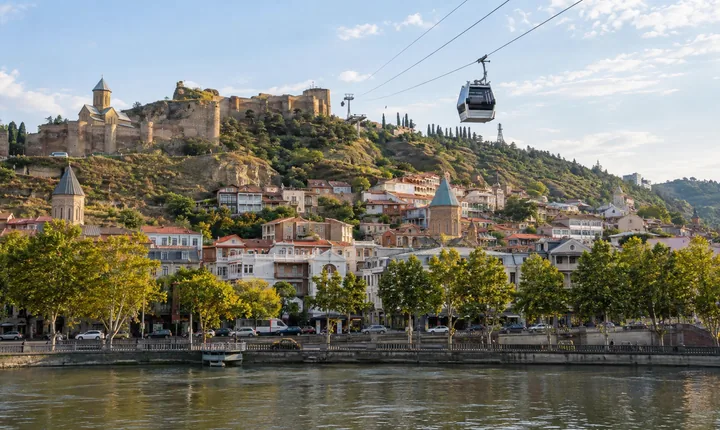 A cable car crosses above Tbilisi's river with hillside houses behind it A cable car crosses above Tbilisi's river with hillside houses behind it
